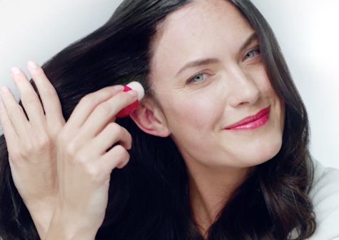 A woman wearing white holds her hair out and applies Garnier Express Retouch to her temple.